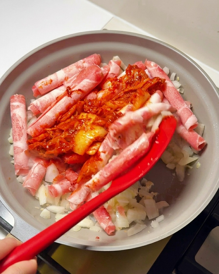 Ein roter Spatel gibt die Kimchi-Chilipaste-Mischung zu den Fleischröllchen und Zwiebeln in die Pfanne.
