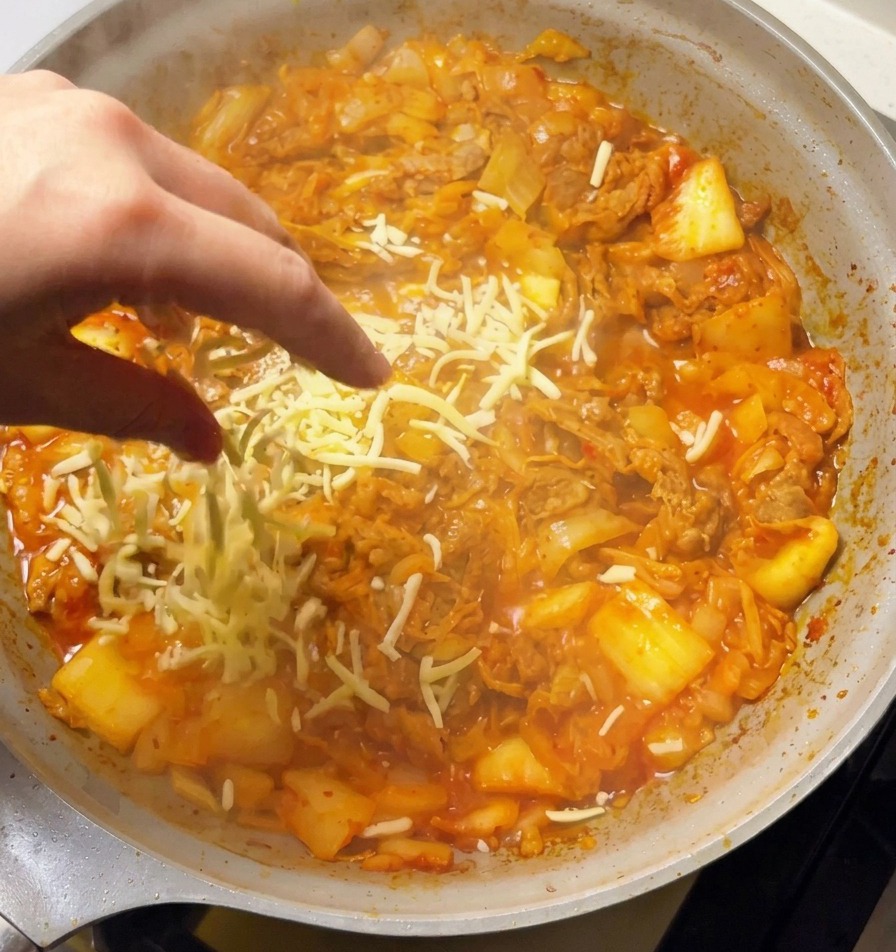 Eine Hand streut reichlich geriebenen weißen Mozzarella über das brutzelnde Kimchi-Rindfleisch-Gemisch.