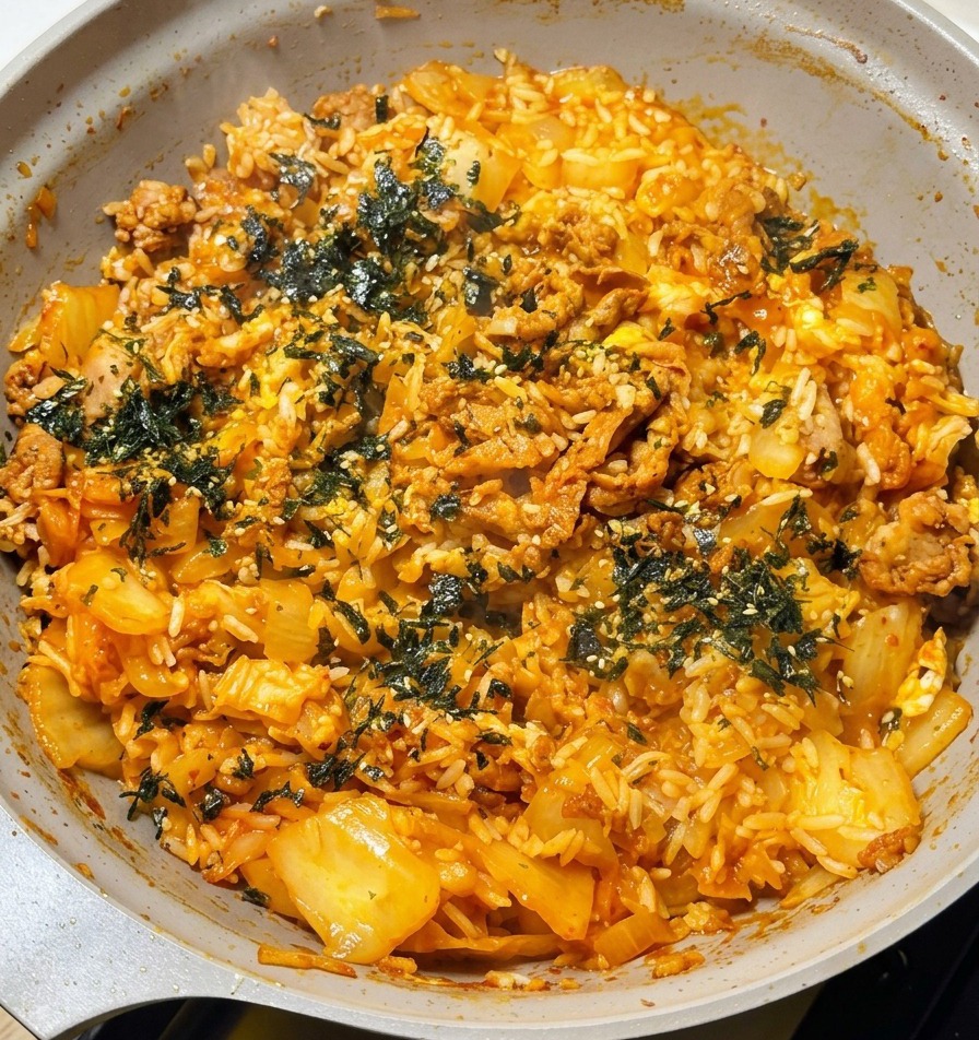Eine Hand streut dunkelgrüne geröstete Algenflocken über den fertigen, dampfenden Kimchi-Reis.
