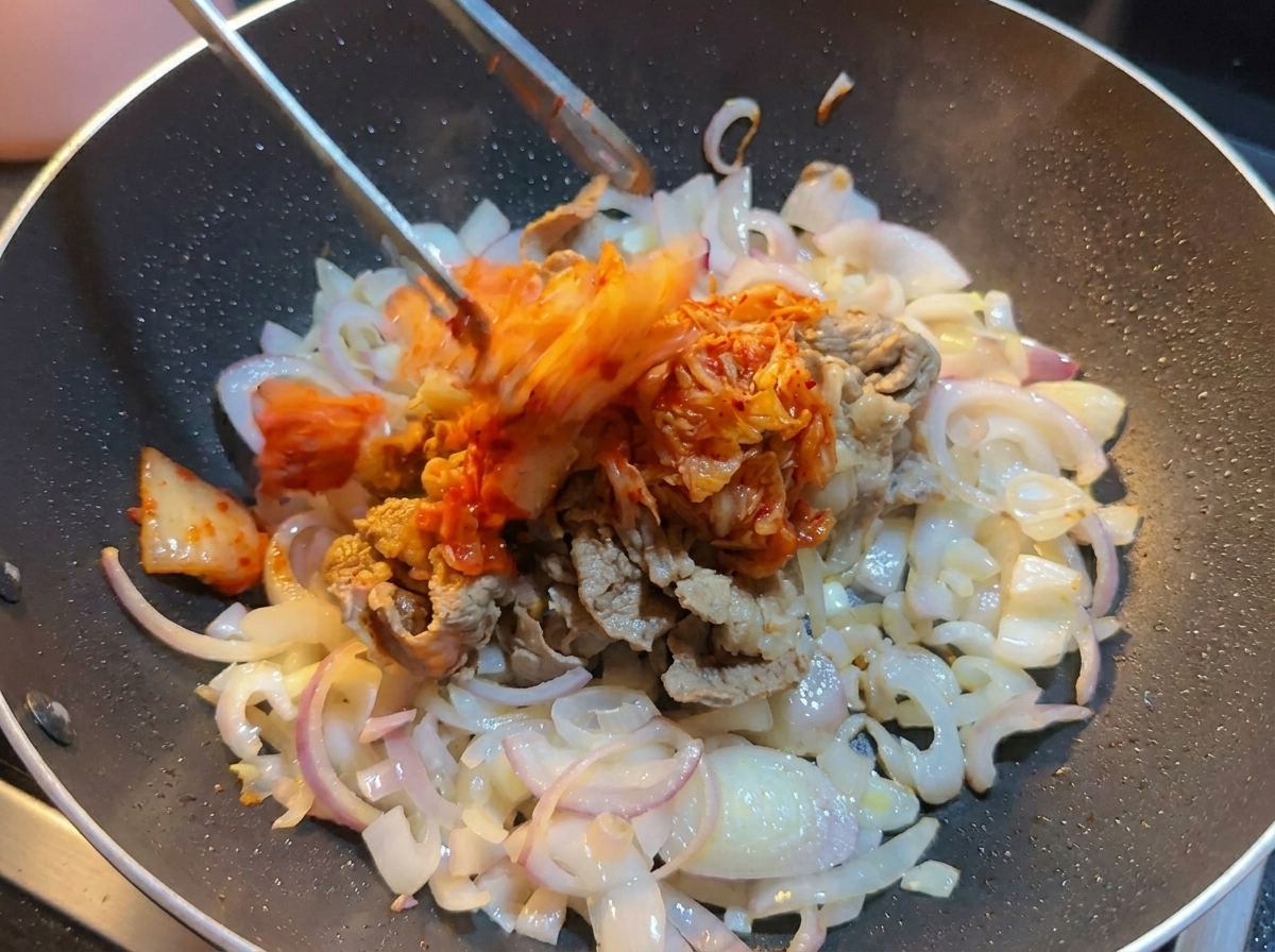 Gehacktes Kohl-Kimchi wird mit einer Zange in einen Wok zu Rindfleisch und Zwiebeln gegeben.