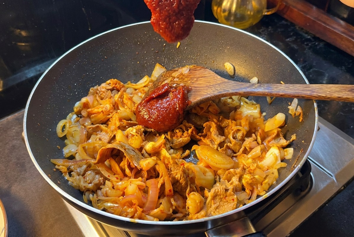 Ein Pfannenwender hält einen großen Löffel rote koreanische Chilipaste über eine Pfanne mit Rindfleisch, Zwiebeln und Kimchi.