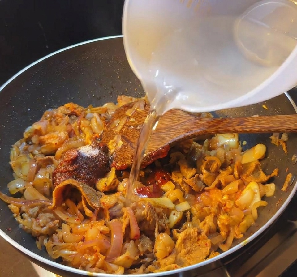 Wasser wird aus einer weißen Schale in eine Pfanne mit Fleisch und scharfer roter Kimchi-Sauce gegossen.