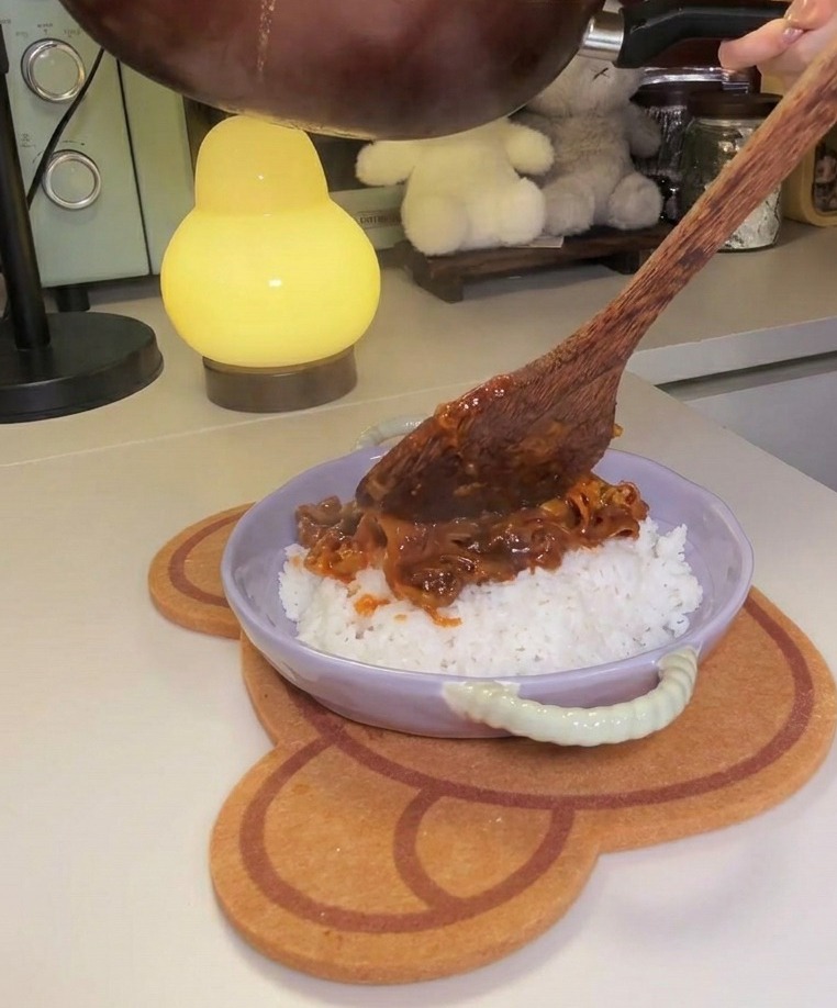 Eine Pfanne wird geneigt, um eine dicke Fleisch-Kimchi-Mischung über eine Portion weißen Reis in einer Schale zu geben.