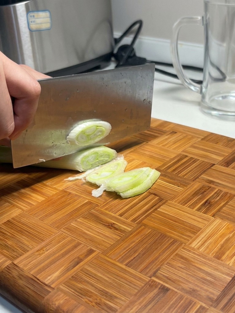 Ein großes Messer schneidet durch einen dicken Lauch auf einem Holzbrett und erzeugt runde Scheiben.