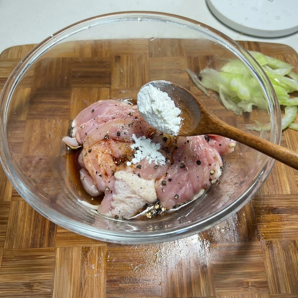 Ein Holzlöffel gibt weiße Maisstärke zu einer Glasschüssel mit gewürzten rohen Hähnchenstücken.