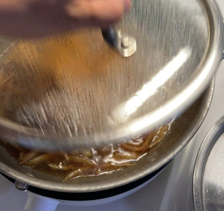 Eine Hand setzt einen Glasdeckel auf eine Pfanne, in der Hähnchen und Zwiebeln in einer dunklen Sauce köcheln.