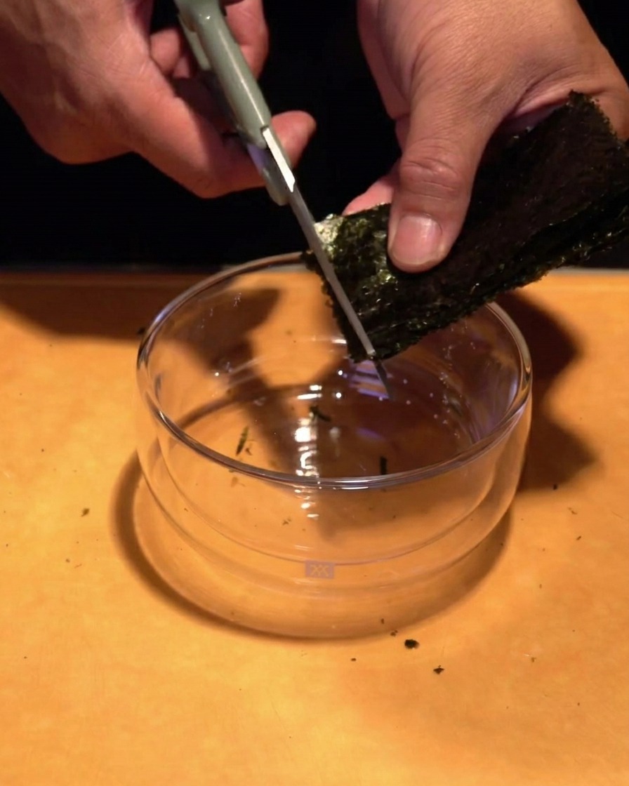 Hände schneiden mit einer Küchenschere getrocknete Nori-Algen in feine Streifen über eine Glasschüssel.