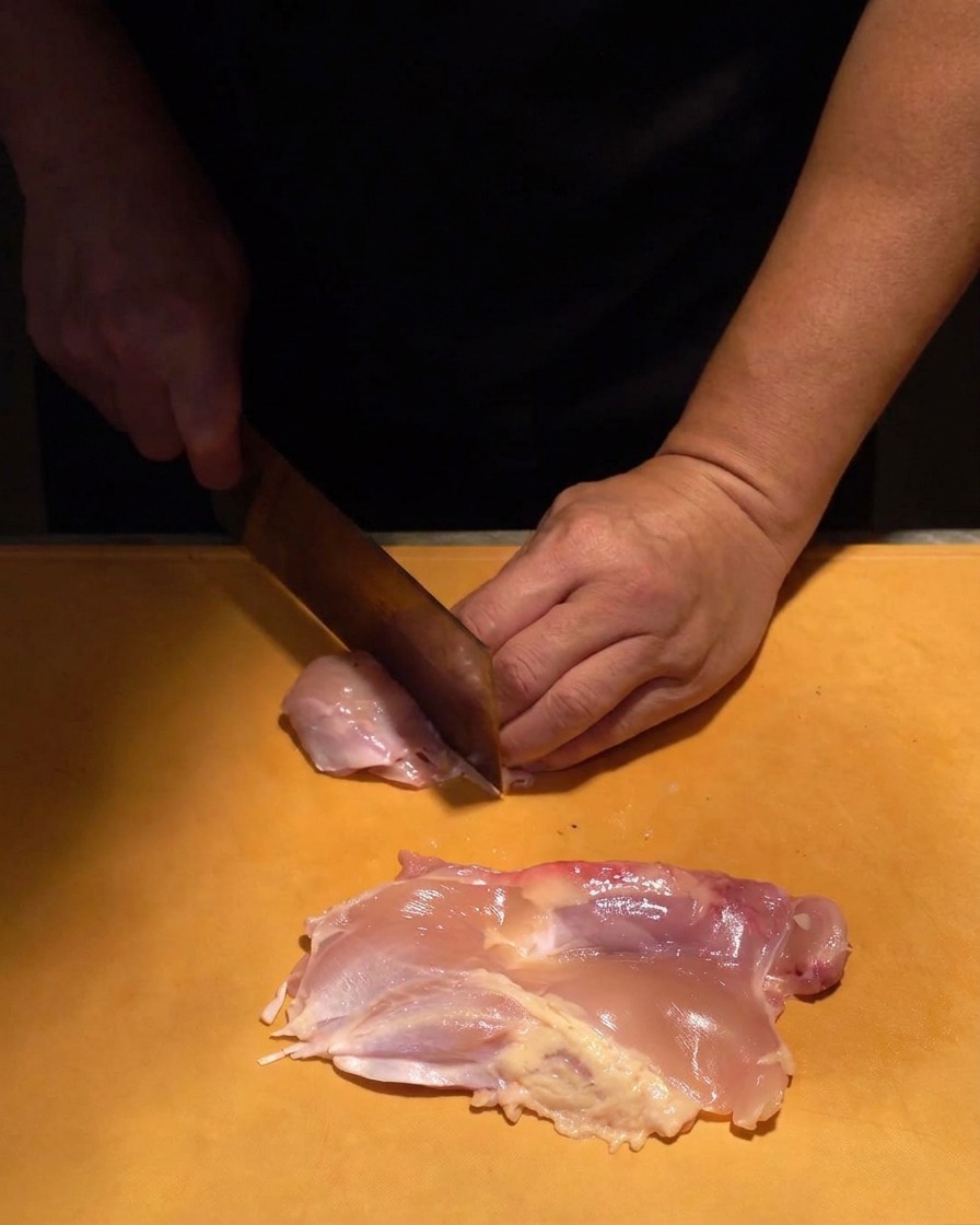 Rohes Hähnchenfleisch wird mit einem scharfen Messer auf einem orangefarbenen Brett in gleichmäßige Stücke gewürfelt.