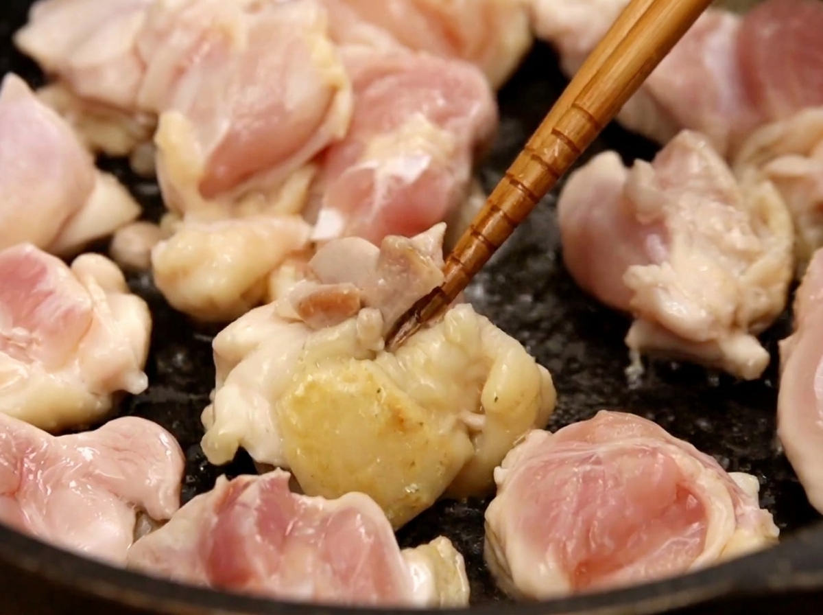 Mundgerechte Hähnchenstücke werden in einer dunklen Pfanne mit Stäbchen angebraten, einige Stellen sind bereits gebräunt.