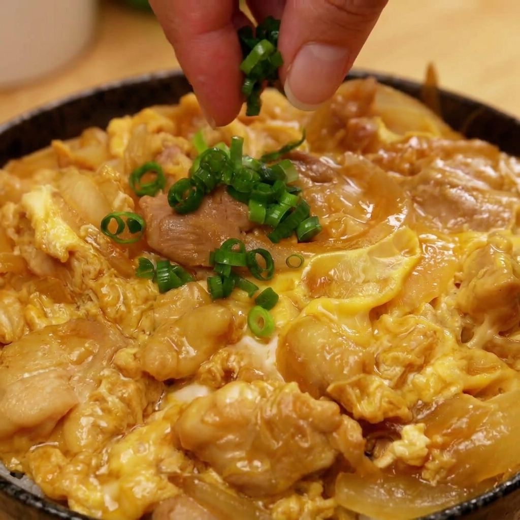 Eine Hand streut gehackte Frühlingszwiebeln über die Mitte der fertigen Oyakodon-Bowl.
