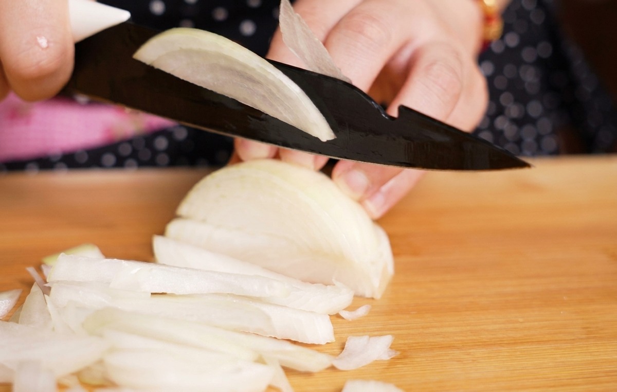 Eine Person schneidet eine weiße Zwiebel mit einem schwarzen Messer in feine Streifen auf einem hellen Holzbrett.