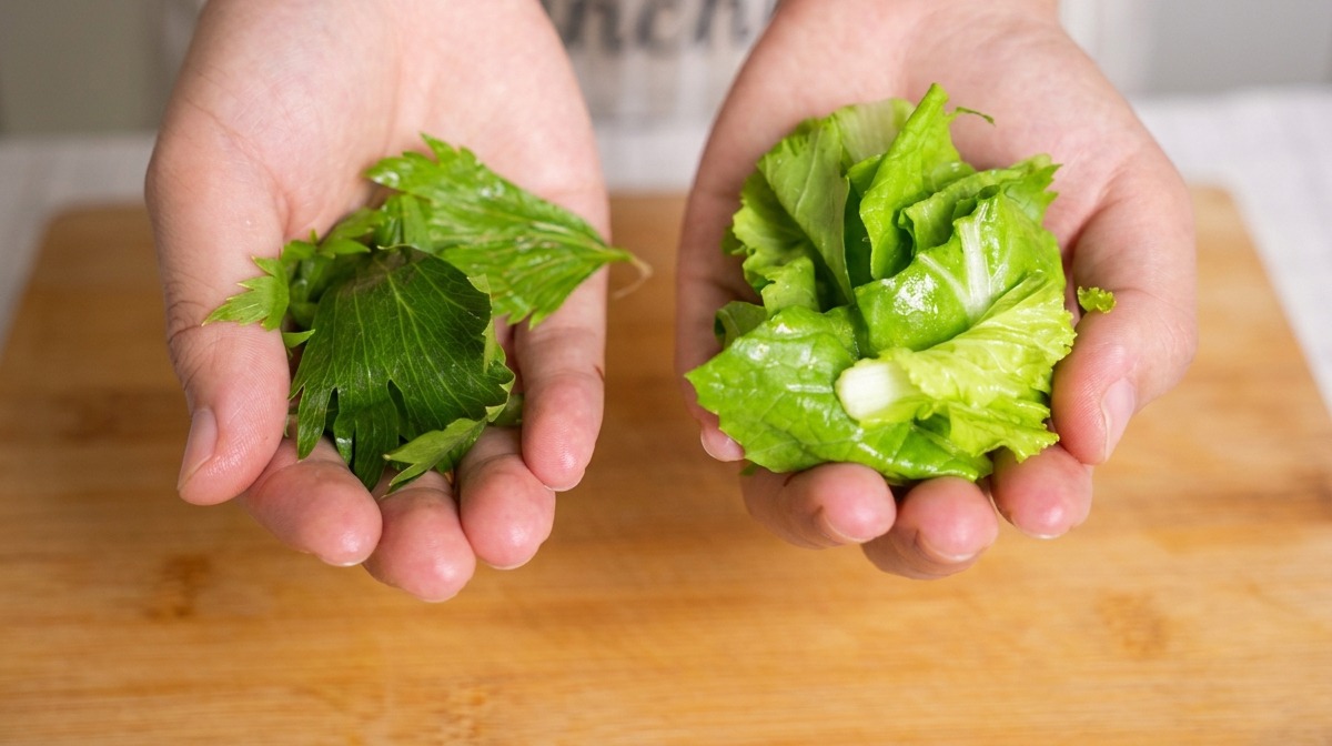 Zwei Hände halten gehackte grüne Sellerieblätter und Baby-Pak-Choi über einem Bambusbrett.