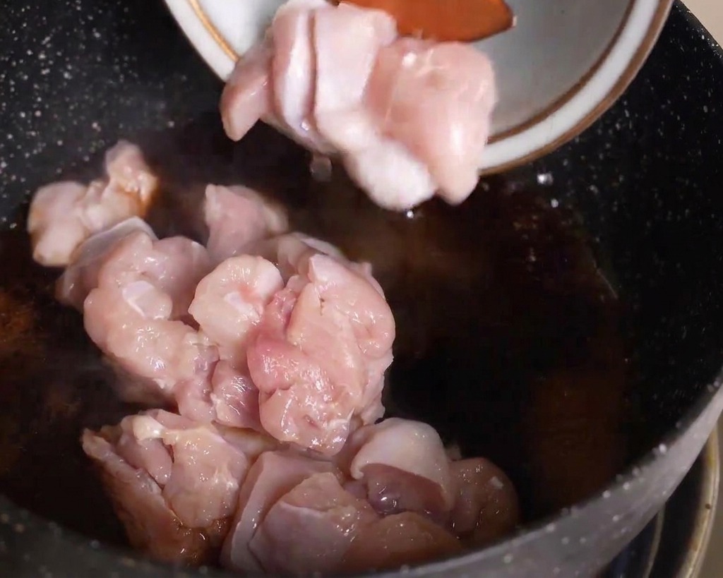 Rohe Hähnchenstücke werden aus einer Schüssel in eine Pfanne mit dunkel köchelnder Sauce gegeben.