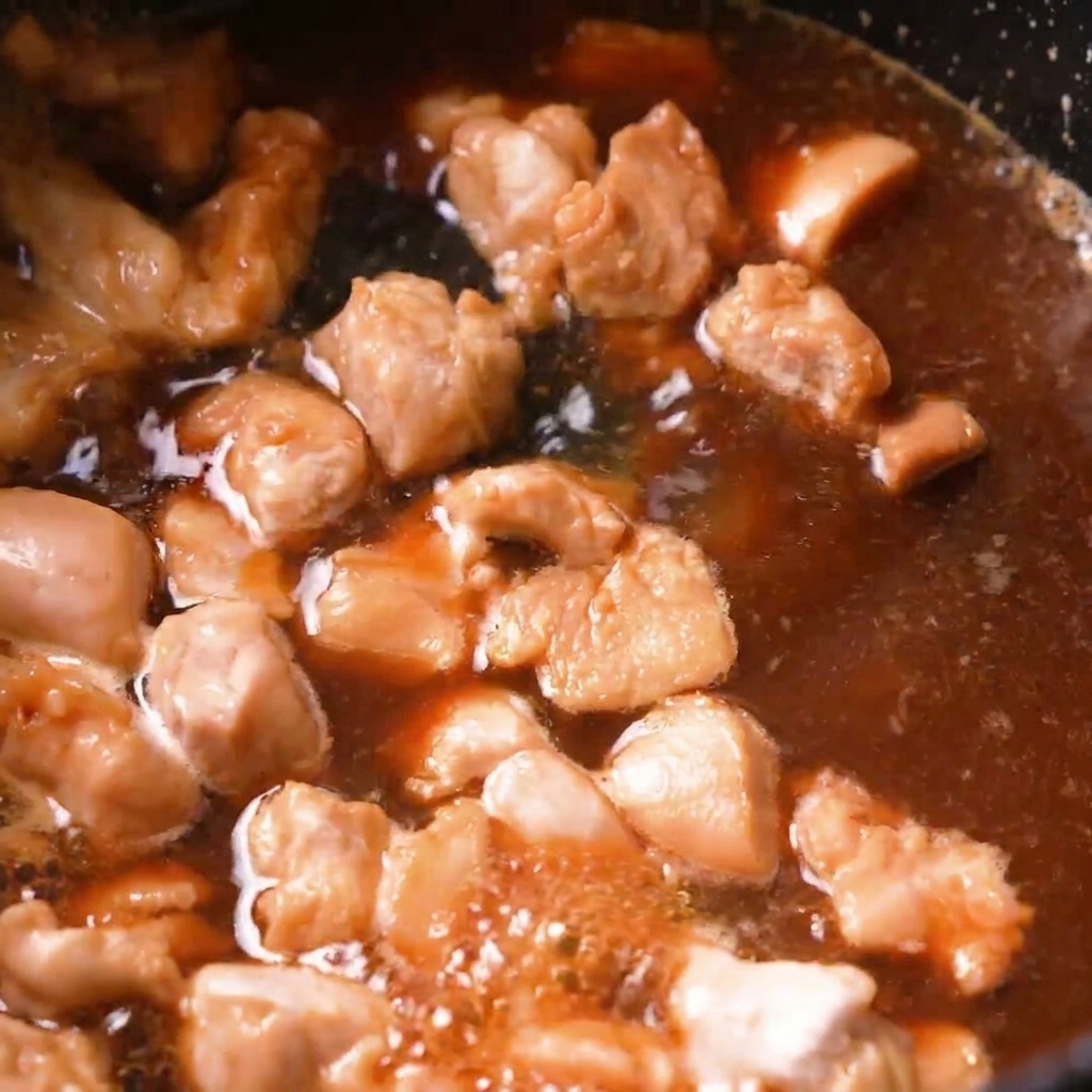 Hähnchenstücke köcheln in einer blubbernden braunen Dashi-Brühe in einer dunklen Pfanne.