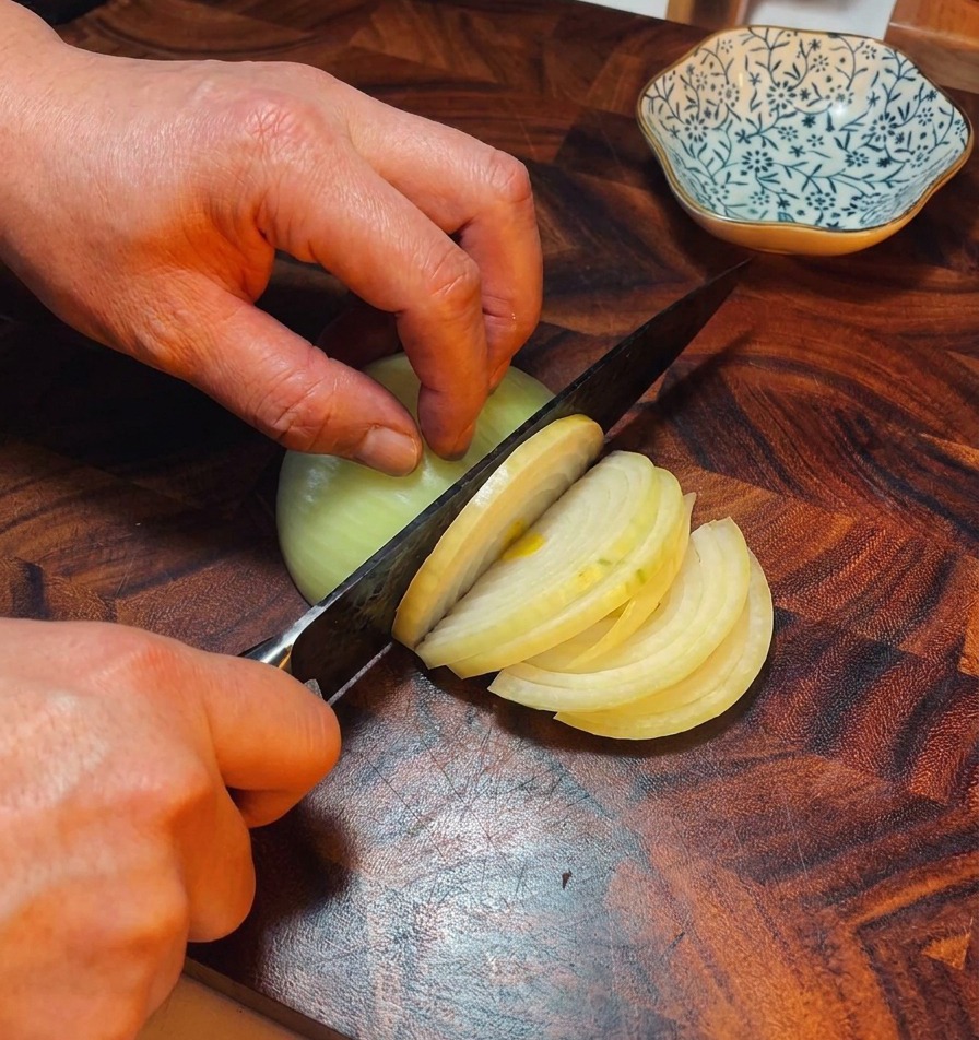 Hände schneiden eine gelbe Zwiebel in feine Streifen auf einem dunklen Holzbrett.