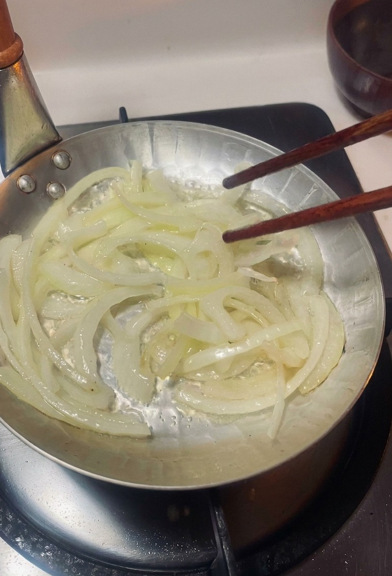 Zwiebelstreifen werden mit Stäbchen in einer kleinen Pfanne über einem Gasbrenner gerührt.