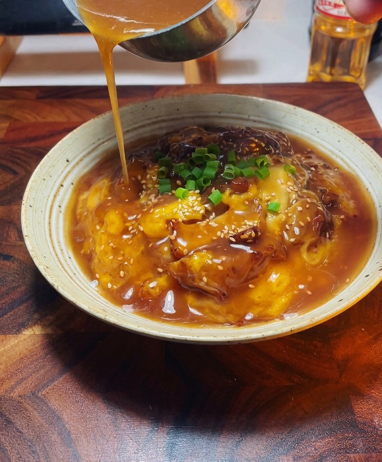 Nahaufnahme einer Hand, die mit einer Kelle Sauce über die angerichtete Oyakodon-Bowl gießt.