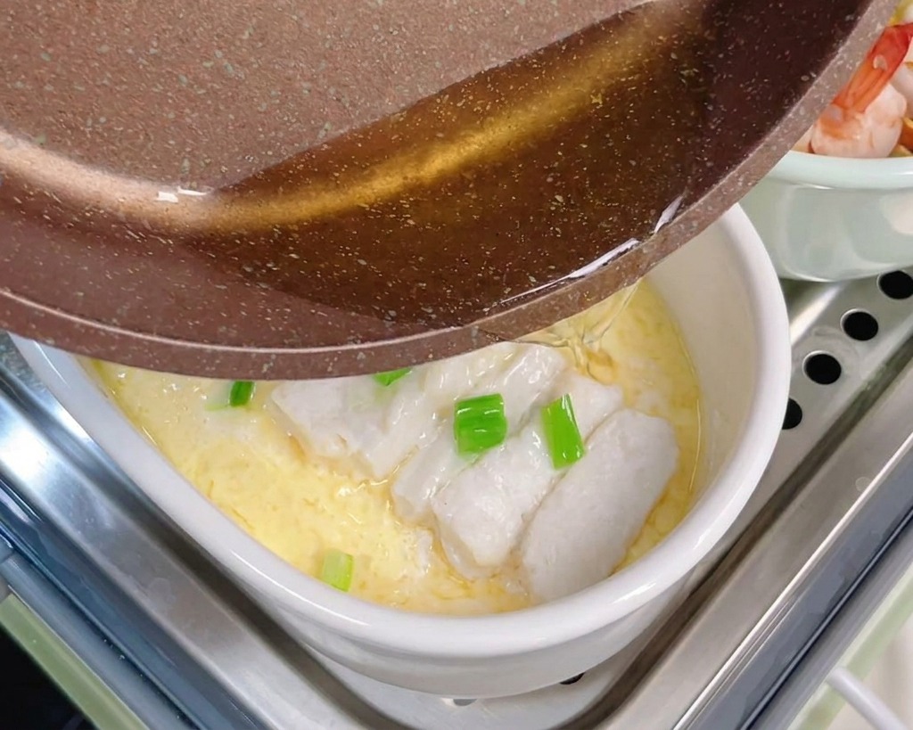 Heißes Öl wird aus einer Pfanne über Frühlingszwiebeln und gedämpften Fisch auf dem Eierstich gegossen.
