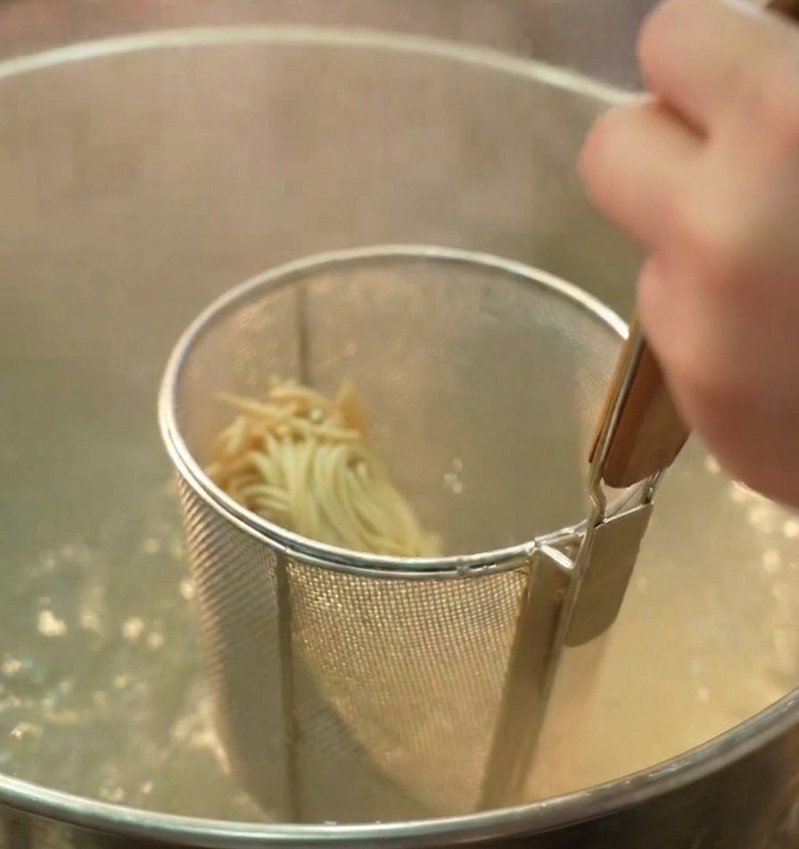 Ein Nudelsieb mit Ramen-Nudeln wird in einen Topf mit sprudelnd kochendem Wasser getaucht.