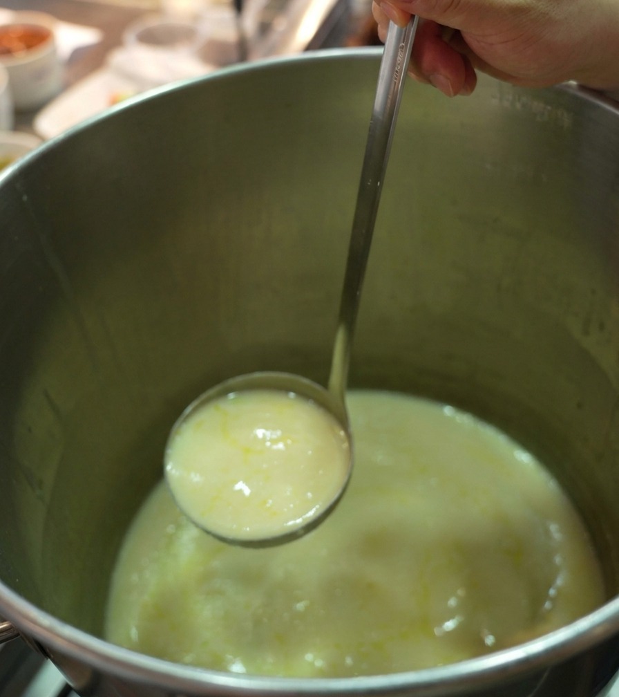 Eine Edelstahlkelle schöpft dicke, cremige gelbe Hühnerbrühe aus einem großen Kochtopf.