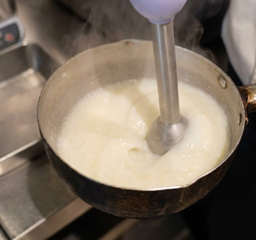 Ein Stabmixer in einem Topf mit gelber Flüssigkeit erzeugt einen dicken, weißen und cremigen Schaum an der Oberfläche.