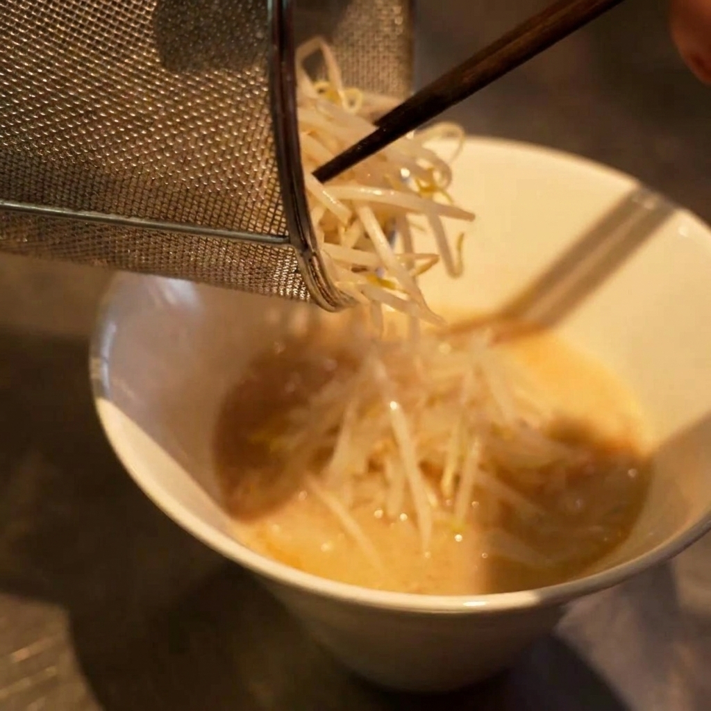 Ein kleines Sieb gibt einen Haufen weißer Sojasprossen auf eine Schüssel mit Ramen und Brühe.