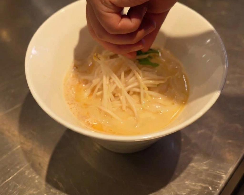 Eine Hand legt vorsichtig frische grüne Spinatblätter auf die Sojasprossen in der Ramen-Schüssel.