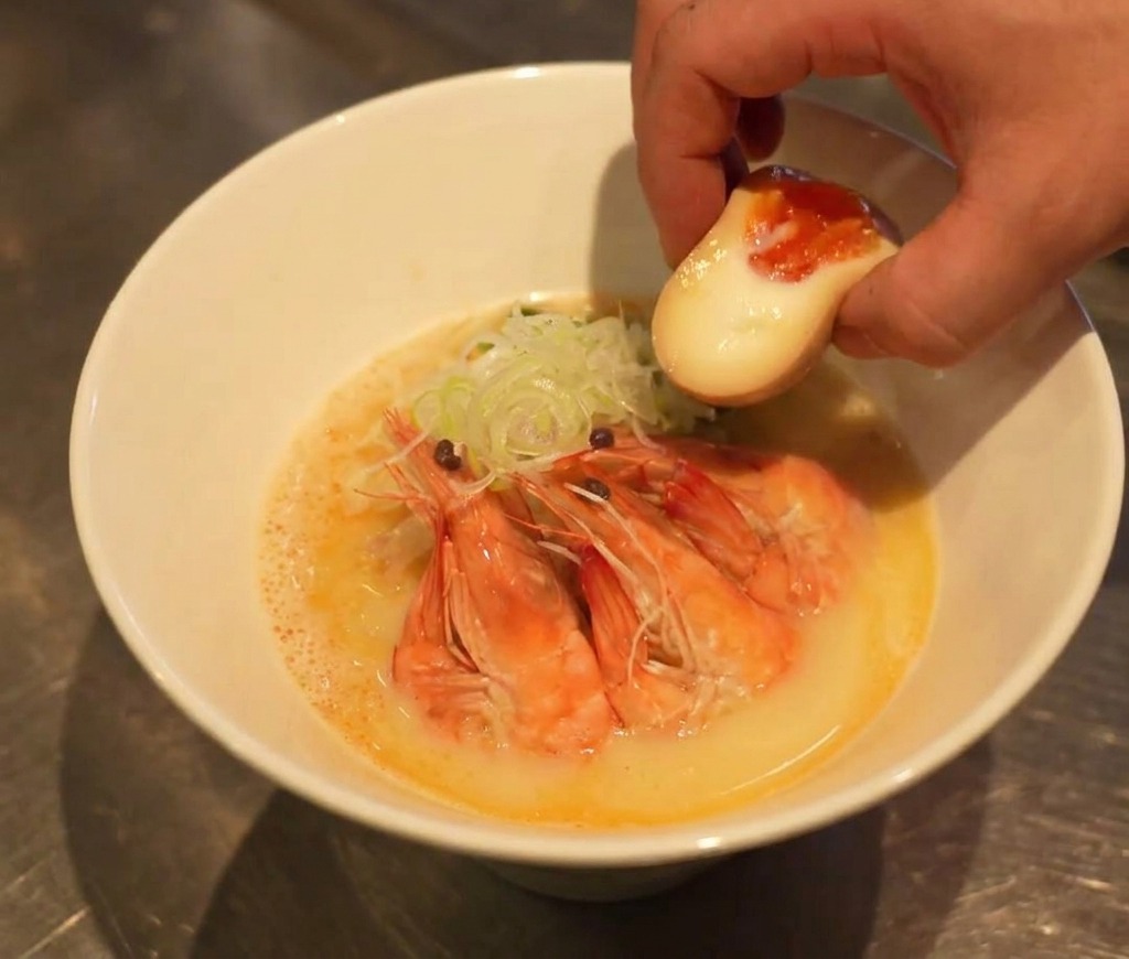 Eine Hand platziert ein halbiertes Ei mit flüssigem, dunklem Dotter in der fast fertigen Ramen-Schüssel.