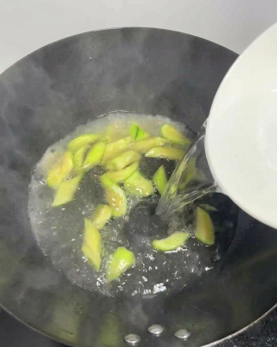 Klares Wasser wird aus einer weißen Schale in den sprudelnden Wok mit Seidenmelonenscheiben gegossen.
