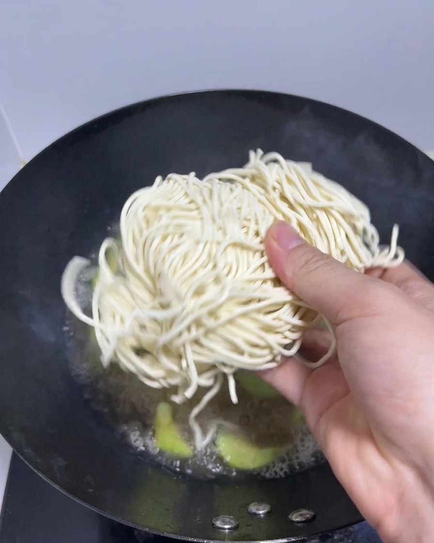 Eine Hand gibt ein Bündel frische Ramen-Nudeln in einen Wok mit kochender Brühe.