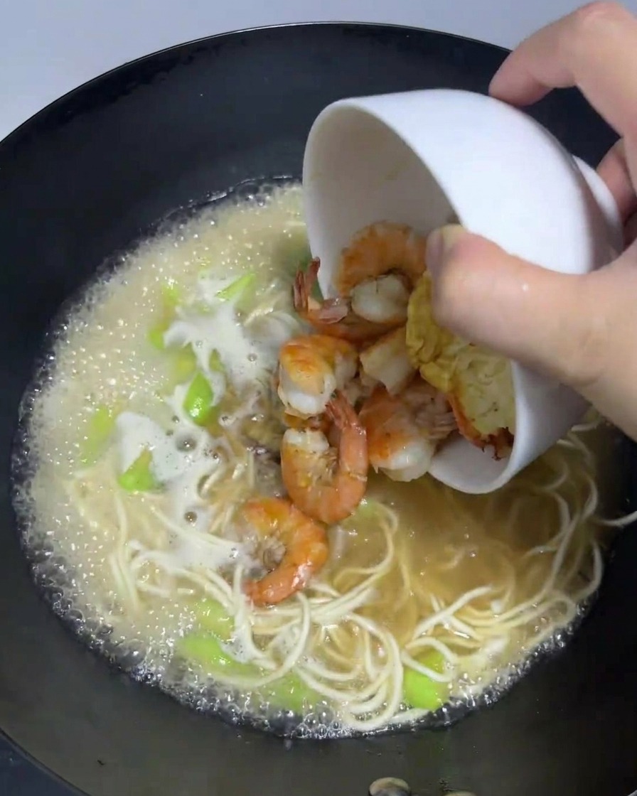 Gekochte Garnelen und ein Spiegelei werden aus einer Schale zurück in den köchelnden Wok mit Ramen-Nudeln gegeben.