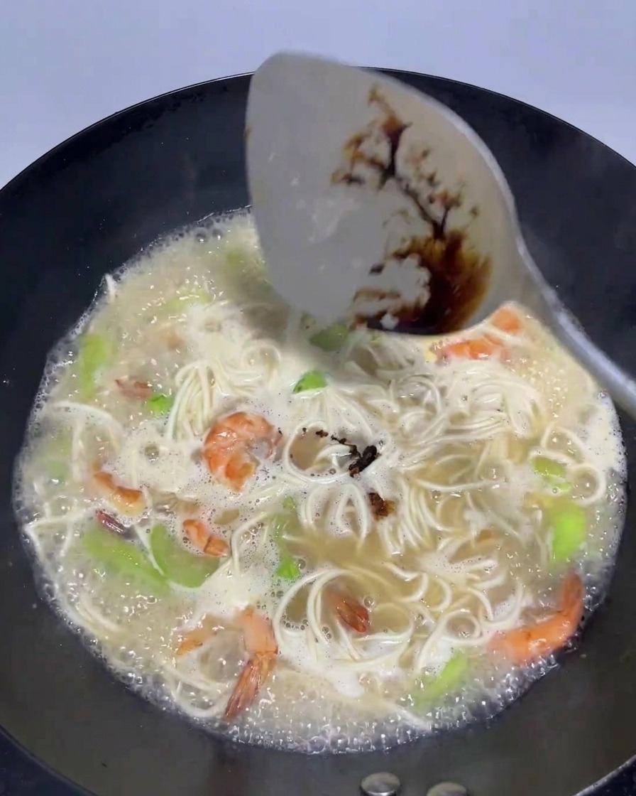 Ein Pfannenwender gießt einen Schuss dunkle Sojasauce in einen Wok voller sprudelnder Brühe, Nudeln und Garnelen.