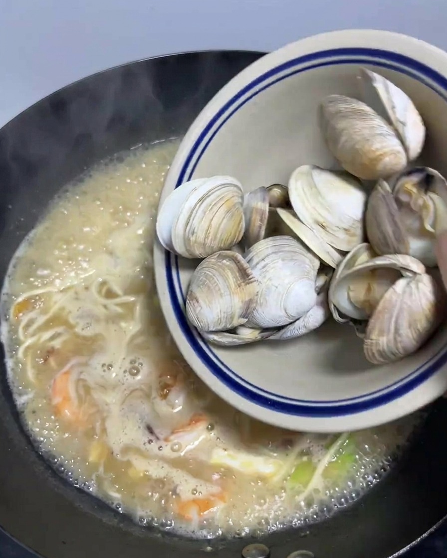 Eine Schale mit blanchierten Venusmuscheln wird in einen großen Wok mit köchelnden Meeresfrüchte-Ramen geschüttet.