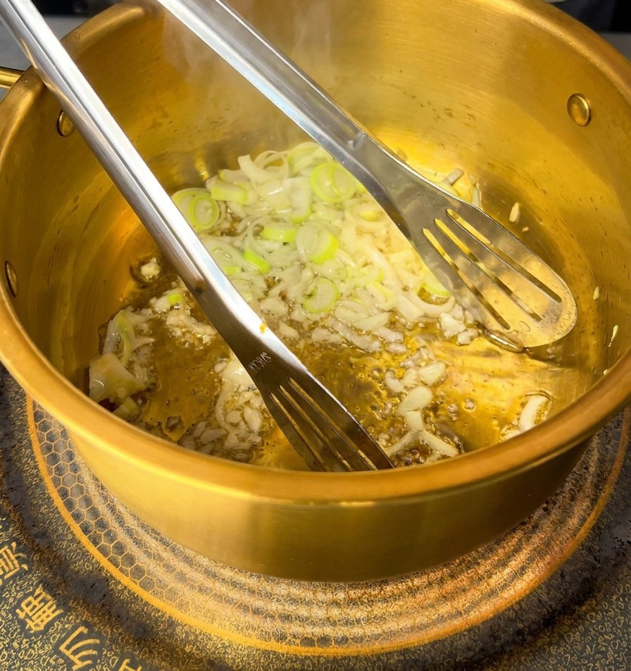 Rohes rosa Hackfleisch wird mit Metallzangen in angebratenen Knoblauch und Zwiebeln in einem goldenen Topf eingerührt.