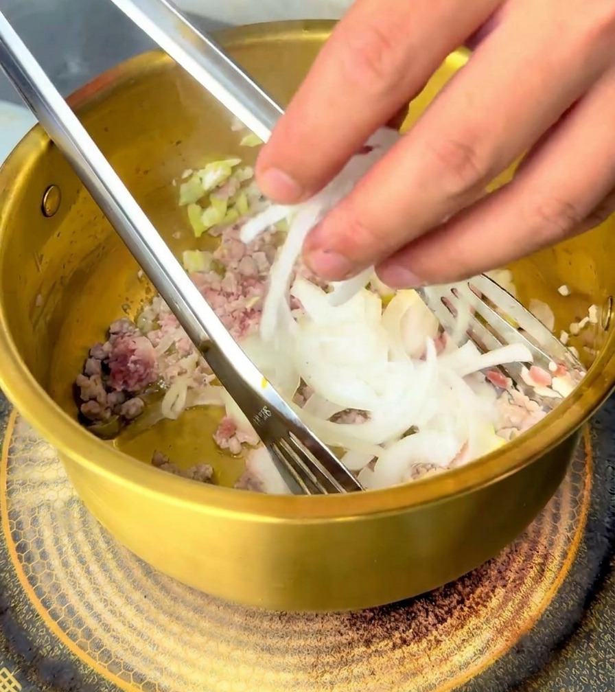 Eine Hand gibt eine Handvoll weißer Zwiebelscheiben in einen goldenen Messingtopf mit gebratenem Fleisch und Aromaten.