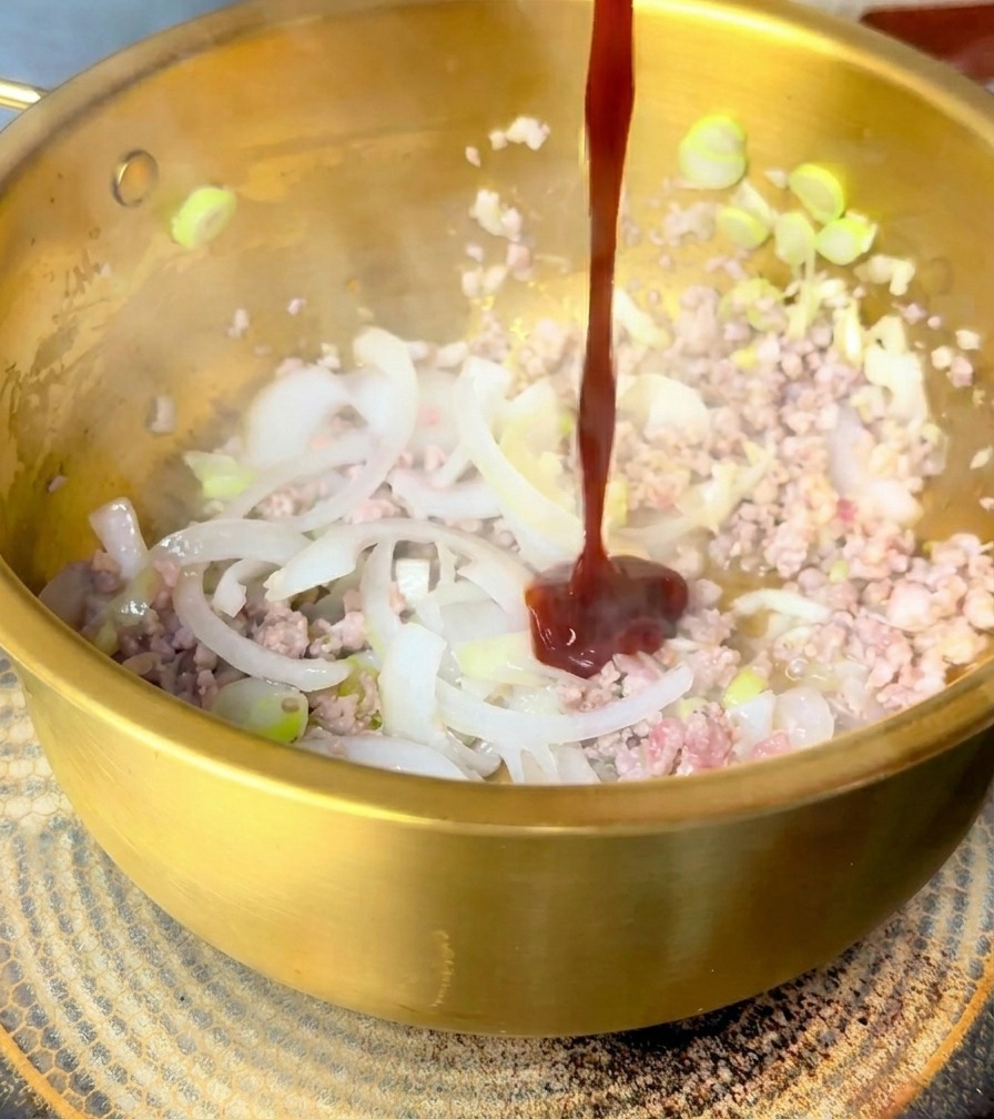 Ein dicker Strahl dunkelroter Sauce wird über gebratenes Schweinefleisch und Zwiebeln in einem goldenen Topf gegossen.