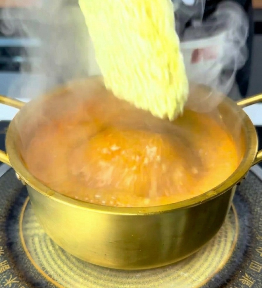 Eine Hand legt einen Block getrocknete Ramen-Nudeln in einen goldenen Topf mit sprudelnder, orangeroter Brühe.