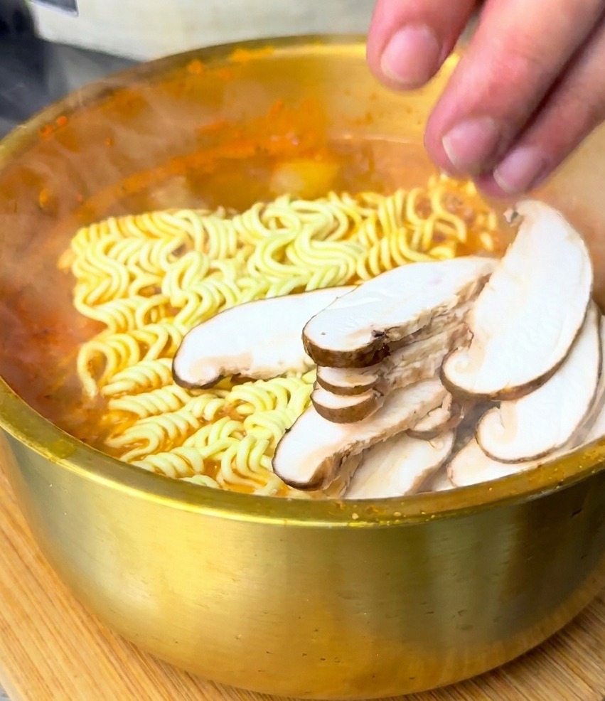 Eine Hand legt geschnittene Shiitake-Pilze auf einen Block Wellennudeln in kochender roter Suppe.