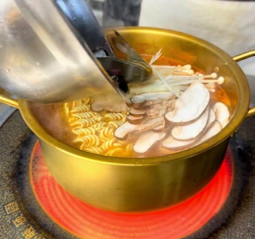 Frische Garnelen und Muscheln werden aus einer Edelstahlschale in einen goldenen Topf mit Nudeln und Pilzen gegeben.