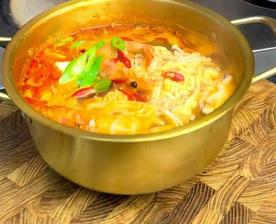 Scharfe Meeresfrüchte-Ramen in einem goldenen Topf, garniert mit frischen Chili-Scheiben, auf einem rustikalen Holzbrett.