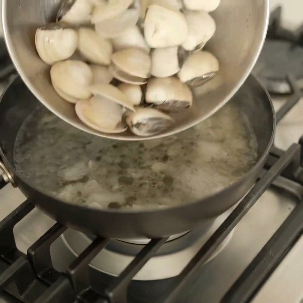 Frische weiße Venusmuscheln werden aus einer Metallschüssel in eine dunkle Pfanne auf dem Herd geschüttet.
