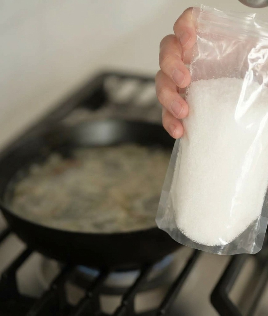 Eine Hand schüttet weißes Würzpulver aus einer Plastikverpackung in die köchelnde Brühe.