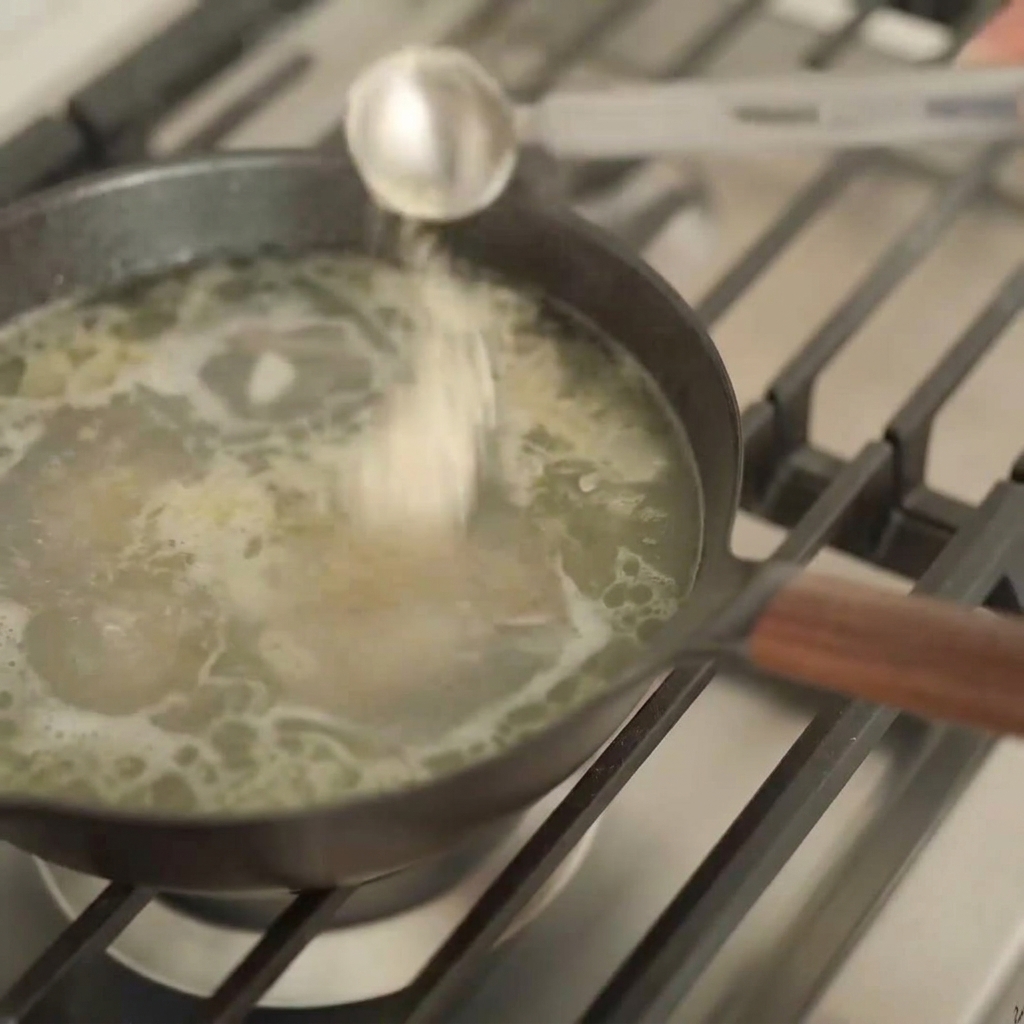 Ein silberner Messlöffel gießt eine dunkle Flüssigkeit in einen Topf mit heller, sprudelnder Ramen-Brühe.