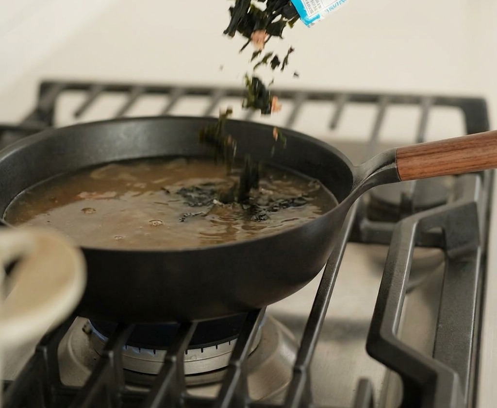 Getrocknete grüne Algenstücke werden in eine Pfanne mit köchelnder, dunkler Brühe gegeben.