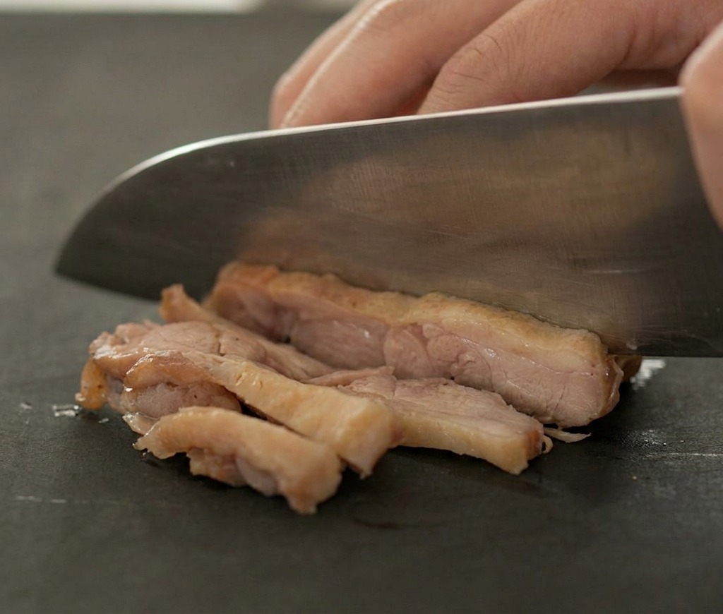 Ein scharfes Messer schneidet gebratenes Entenfleisch auf einem dunklen Brett in Scheiben.