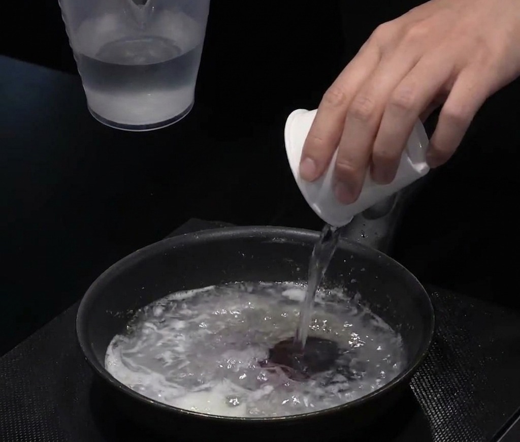 Wasser wird aus einem weißen Becher in eine Pfanne mit der Mehl-Öl-Mischung gegossen.