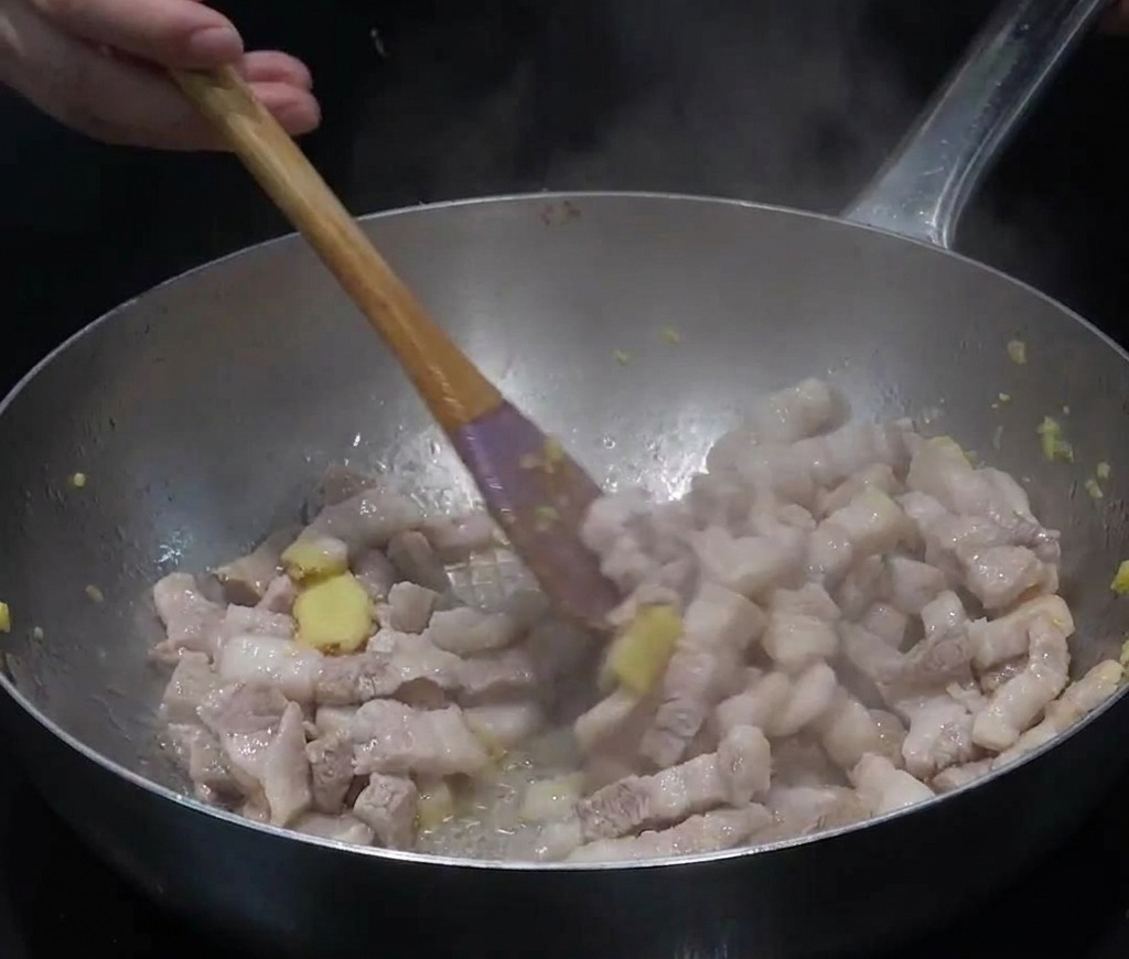 Schweinebauchscheiben und gehackter Ingwer werden in einer Edelstahlpfanne angebraten, es steigt Dampf auf.