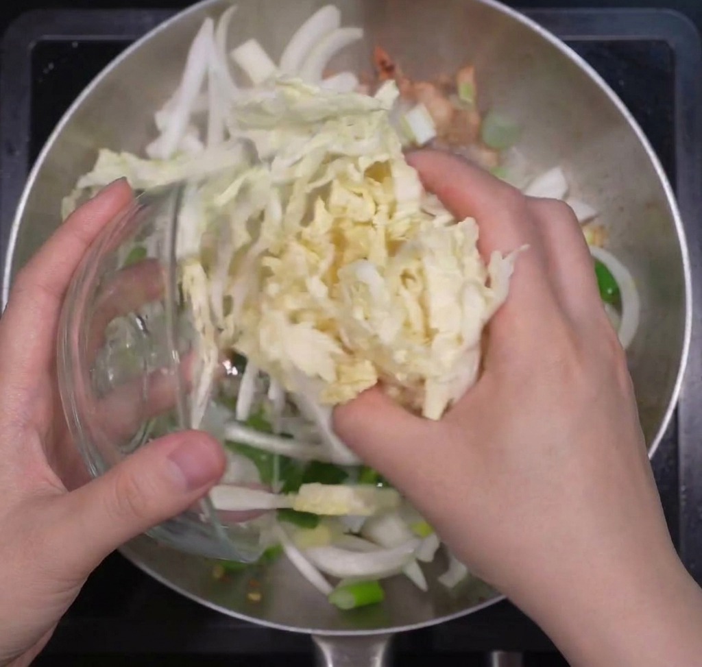 Eine Hand gibt eine große Portion geschnittenen Chinakohl in eine Pfanne zu Zwiebeln und Frühlingszwiebeln.