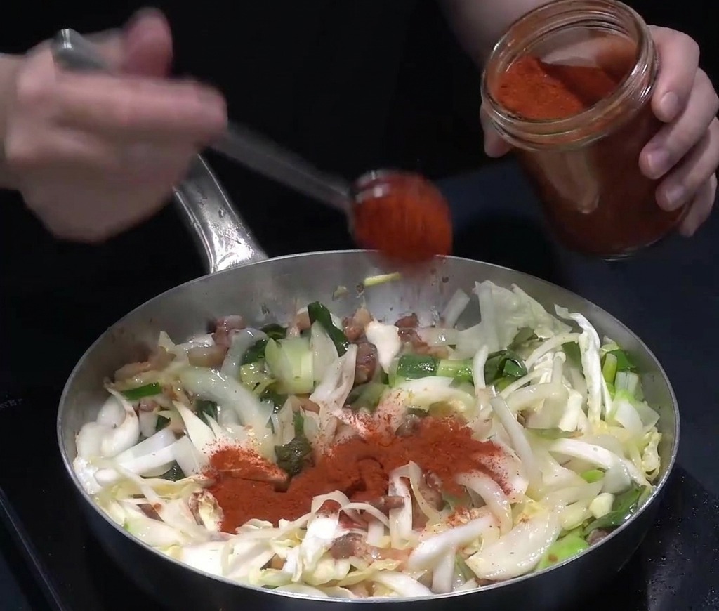 Leuchtend rotes Chilipulver wird mit einem Löffel auf das angebratene Gemüse und Fleisch in der Pfanne gegeben.