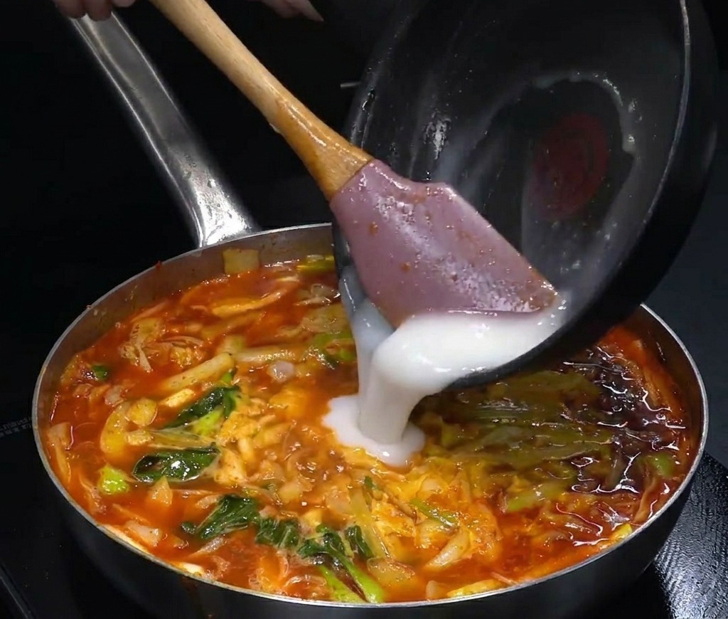 Eine weiße, undurchsichtige Flüssigkeit wird in einen großen Topf mit brodelnder roter Suppe gegossen.